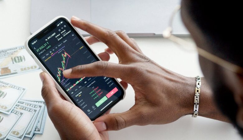 A person trading stocks using a smartphone, showcasing financial data on mobile and laptop screens.